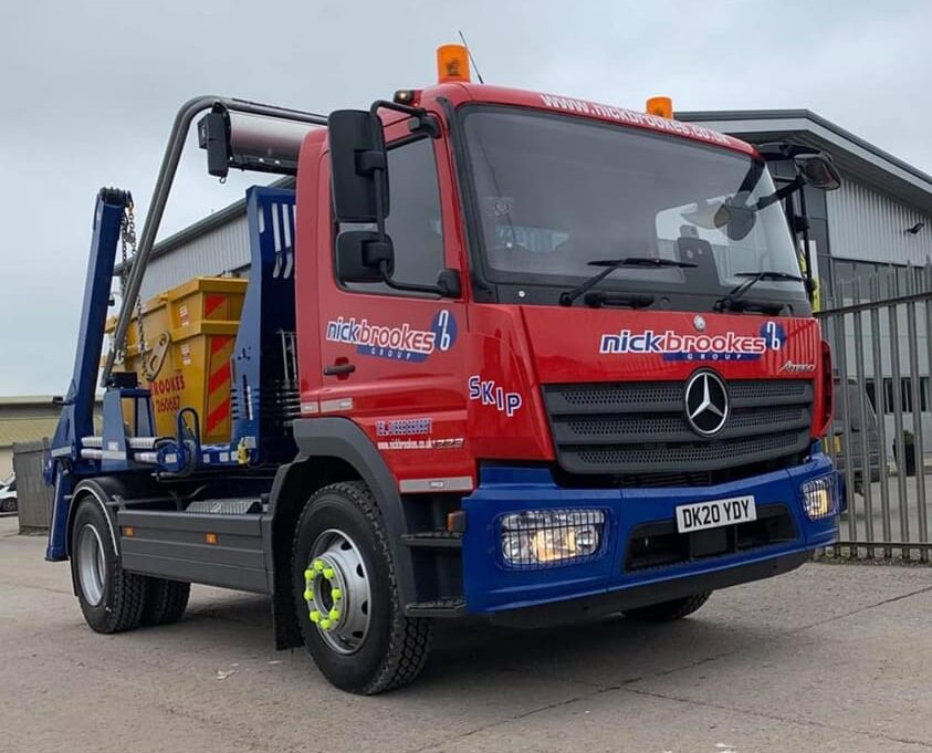 A red and blue Nick Brookes Mercedes skip lorry carrying a yellow skip, parked outside an industrial building.