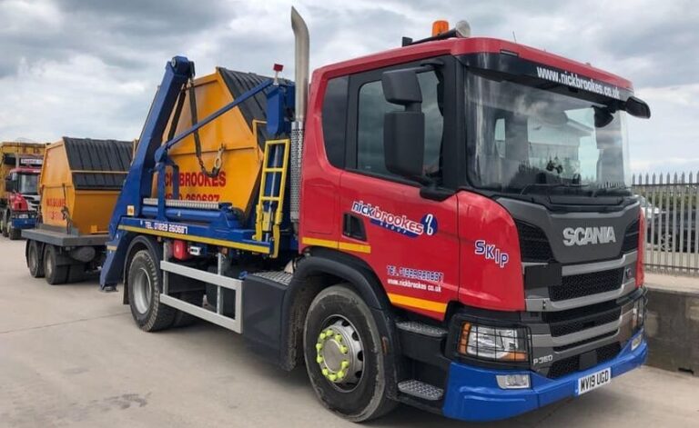 A Nick Brookes skip lorry carrying a yellow skip, with the red and blue branded Scania truck parked on a yard.