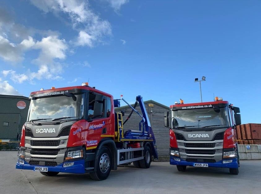 Two red and blue Nick Brookes Scania skip lorries parked side by side on a yard under a bright blue sky.