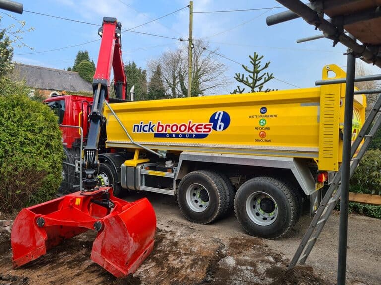 A yellow Nick Brookes Group grab lorry with a red hydraulic grab arm loading waste on a residential site, parked beside trees and houses.